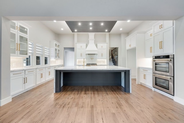 kitchen with decorative backsplash, light wood-type flooring, a large island with sink, white cabinetry, and double oven