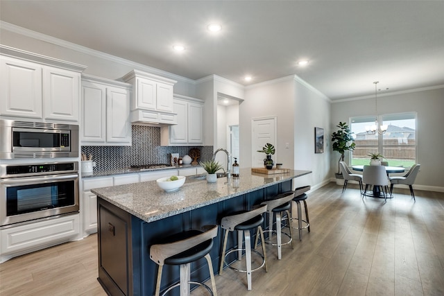 kitchen featuring a center island with sink, stainless steel appliances, light stone countertops, a notable chandelier, and white cabinetry