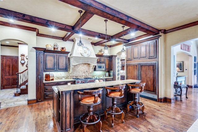 kitchen with coffered ceiling, a center island with sink, decorative backsplash, stainless steel microwave, and dark brown cabinets