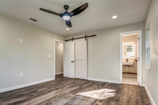 unfurnished bedroom featuring ensuite bathroom, ceiling fan, a barn door, and dark wood-type flooring