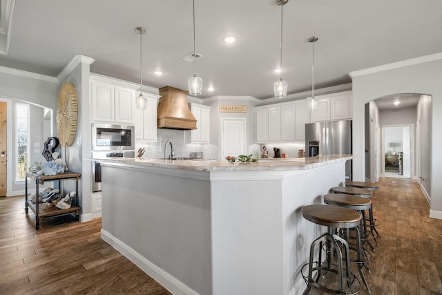 kitchen featuring a kitchen island with sink, stainless steel appliances, custom range hood, pendant lighting, and white cabinetry