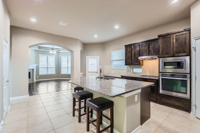 kitchen featuring a center island with sink, appliances with stainless steel finishes, light stone countertops, dark brown cabinetry, and light tile patterned flooring