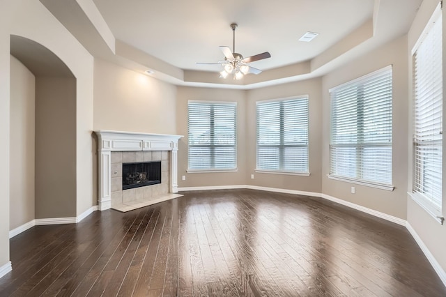 unfurnished living room featuring a raised ceiling, a tiled fireplace, ceiling fan, and dark wood-type flooring