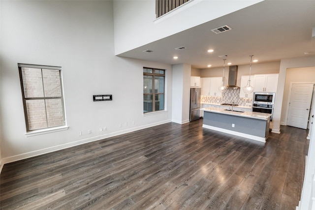 kitchen featuring stainless steel appliances, an island with sink, pendant lighting, white cabinetry, and wall chimney range hood