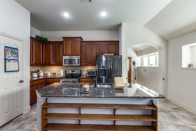 kitchen with stainless steel appliances, dark stone countertops, backsplash, and an island with sink