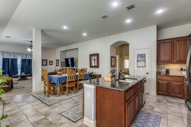 kitchen featuring dishwasher, a kitchen island with sink, dark stone countertops, ceiling fan, and sink