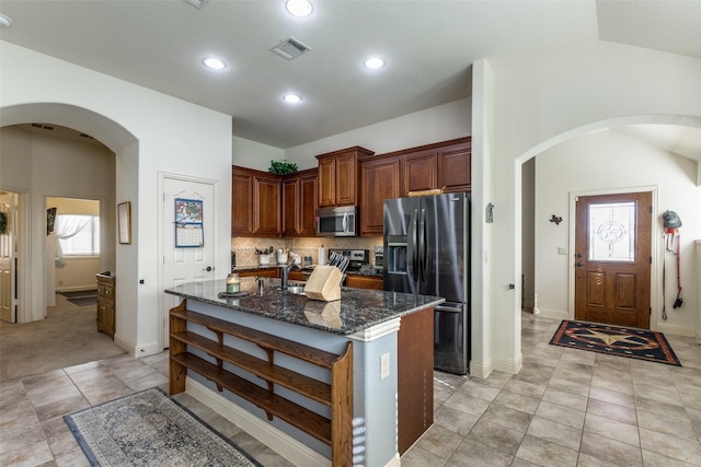 kitchen featuring stainless steel appliances, vaulted ceiling, backsplash, dark stone counters, and a kitchen island with sink