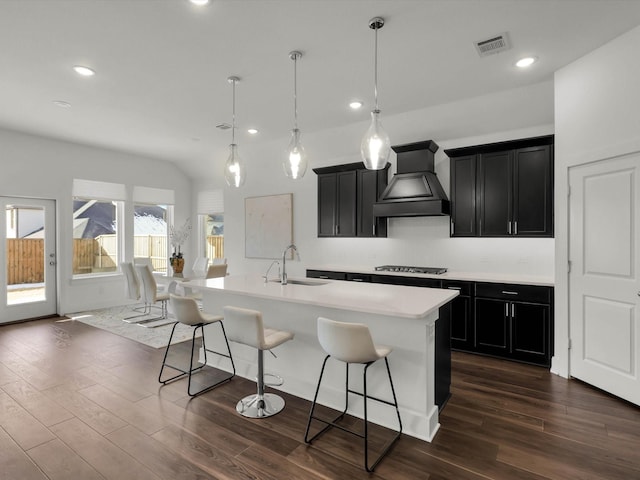 kitchen featuring an island with sink, premium range hood, stainless steel gas stovetop, sink, and decorative light fixtures