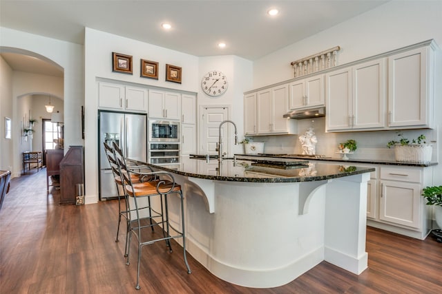 kitchen with built in microwave, white cabinets, dark stone counters, a kitchen island with sink, and stainless steel refrigerator with ice dispenser