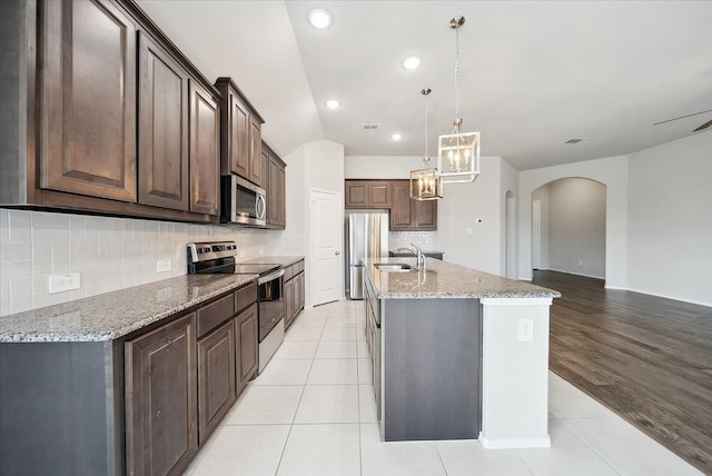 kitchen with sink, light stone counters, an island with sink, dark brown cabinetry, and appliances with stainless steel finishes