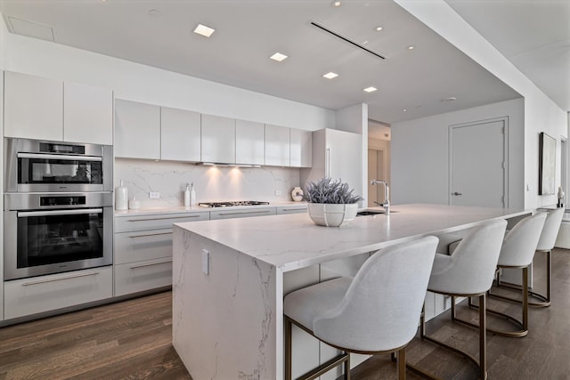 kitchen featuring stainless steel appliances, white cabinets, tasteful backsplash, a center island with sink, and dark hardwood / wood-style flooring