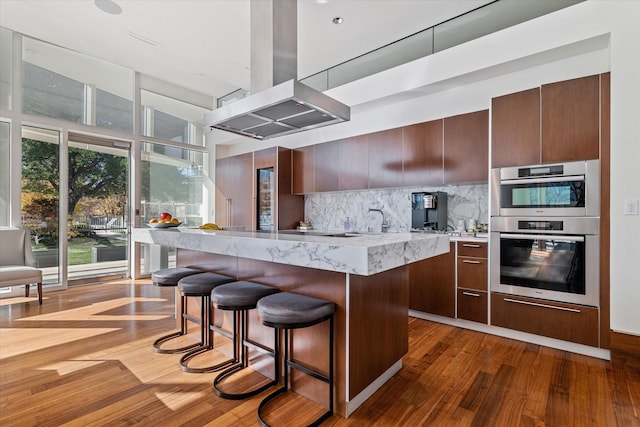 kitchen with island range hood, double oven, dark wood-type flooring, a kitchen island, and tasteful backsplash