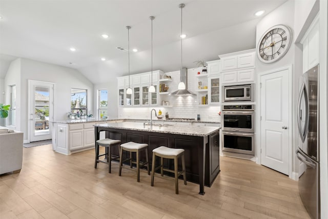 kitchen with wall chimney exhaust hood, a center island with sink, a breakfast bar, white cabinets, and appliances with stainless steel finishes