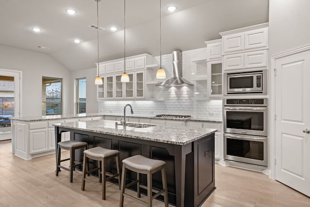 kitchen with wall chimney range hood, sink, appliances with stainless steel finishes, a kitchen island with sink, and white cabinetry