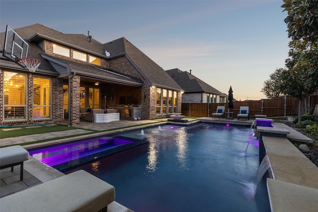 pool at dusk featuring a patio, pool water feature, an in ground hot tub, and area for grilling