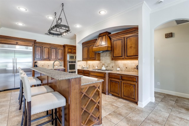 kitchen with custom exhaust hood, built in appliances, backsplash, a kitchen island with sink, and crown molding