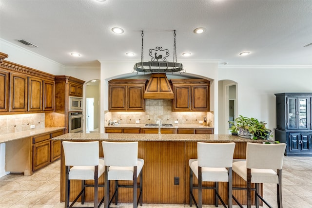 kitchen with premium range hood, an island with sink, appliances with stainless steel finishes, decorative backsplash, and light stone counters