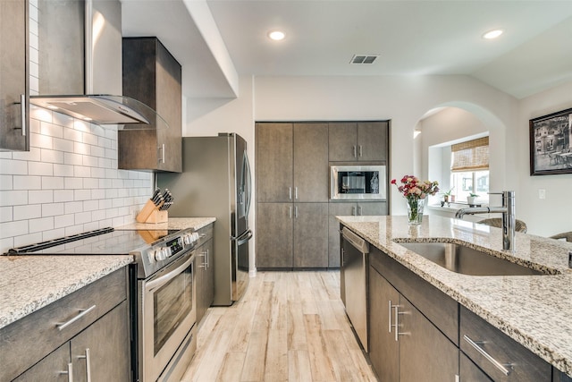 kitchen with wall chimney range hood, sink, light stone countertops, stainless steel appliances, and dark brown cabinets