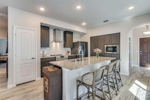 kitchen with light hardwood / wood-style floors, a center island with sink, appliances with stainless steel finishes, wall chimney exhaust hood, and sink