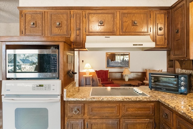 kitchen featuring black electric stovetop, under cabinet range hood, white oven, brown cabinetry, and stainless steel microwave