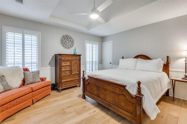 bedroom with ceiling fan, a textured ceiling, light wood-type flooring, and a tray ceiling