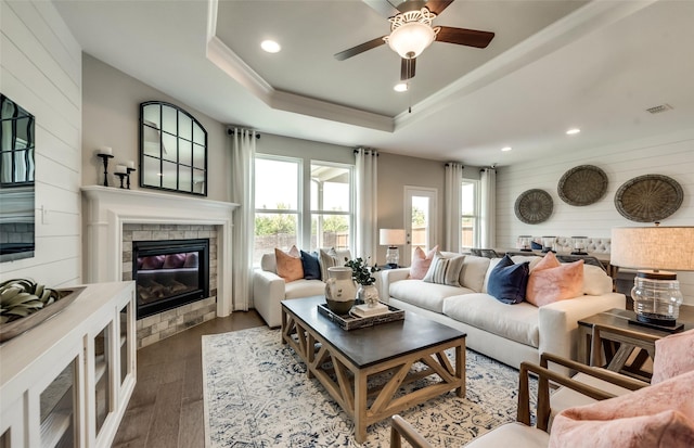 living room with wood-type flooring, wood walls, ceiling fan, a tray ceiling, and a tiled fireplace