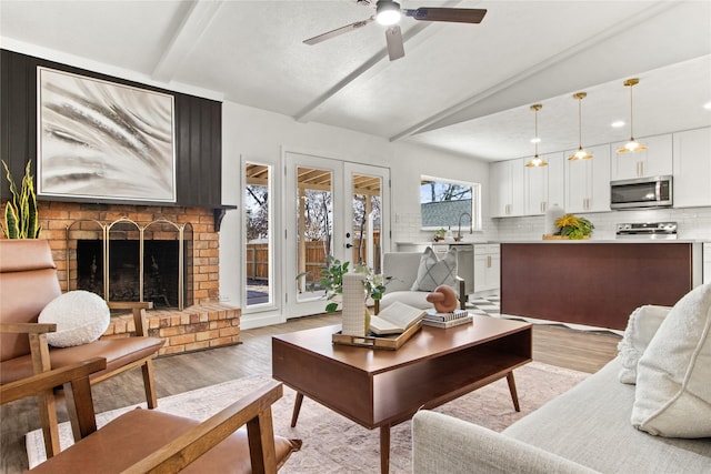 living room featuring ceiling fan, a fireplace, light hardwood / wood-style floors, lofted ceiling with beams, and french doors