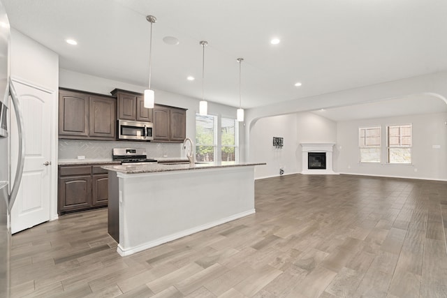 kitchen with pendant lighting, a kitchen island with sink, appliances with stainless steel finishes, dark brown cabinets, and light stone counters