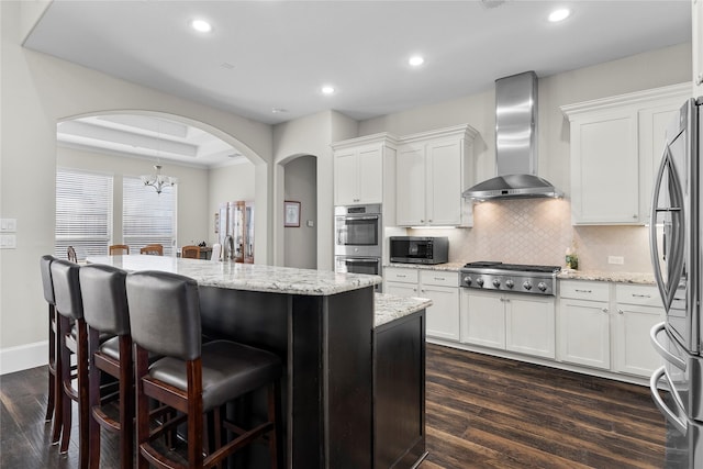 kitchen featuring arched walkways, dark wood-style flooring, a raised ceiling, appliances with stainless steel finishes, and wall chimney range hood