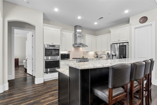 kitchen with white cabinetry, wall chimney range hood, stainless steel appliances, and a center island with sink