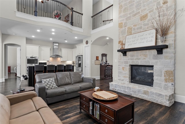 living room featuring arched walkways, a fireplace, a towering ceiling, dark wood-type flooring, and baseboards