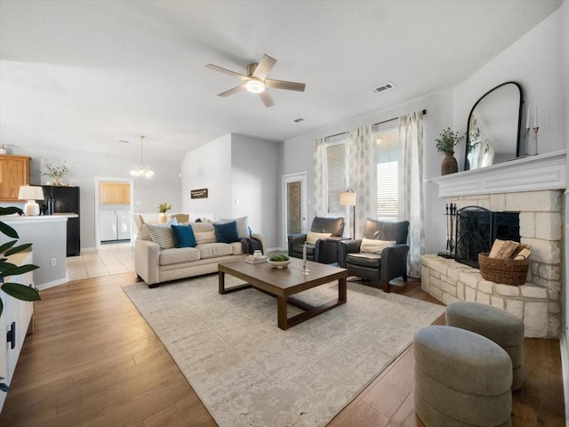 living room featuring light hardwood / wood-style floors, washing machine and clothes dryer, a stone fireplace, and ceiling fan with notable chandelier