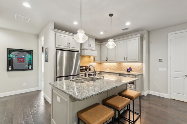 kitchen with a center island with sink, sink, hanging light fixtures, light stone countertops, and stainless steel fridge