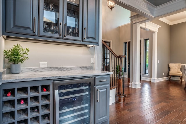 bar with ornate columns, dark wood-type flooring, beverage cooler, crown molding, and light stone counters