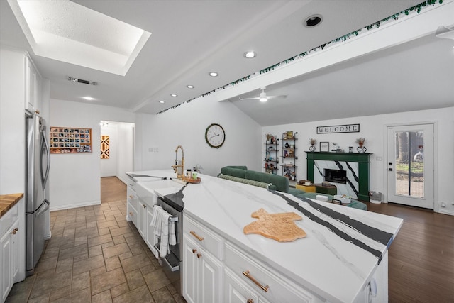 kitchen featuring ceiling fan, lofted ceiling with skylight, a center island with sink, white cabinets, and stainless steel fridge