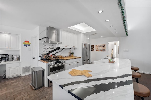 kitchen featuring appliances with stainless steel finishes, wall chimney range hood, white cabinetry, and a center island with sink