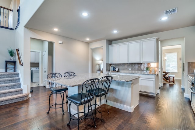 kitchen featuring light stone countertops, white cabinets, a kitchen bar, an island with sink, and backsplash