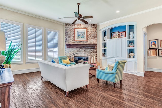 living room with dark wood-type flooring, a brick fireplace, built in features, ornamental molding, and ceiling fan