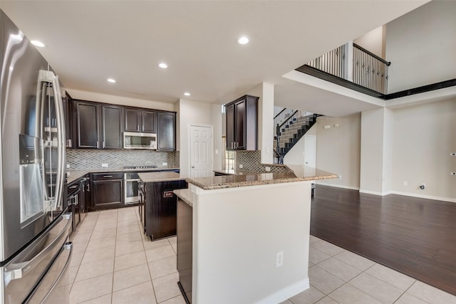 kitchen with a kitchen island, dark brown cabinetry, light stone countertops, stainless steel appliances, and light tile patterned floors