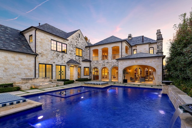 back house at dusk with pool water feature, a pool with hot tub, a patio area, and a balcony