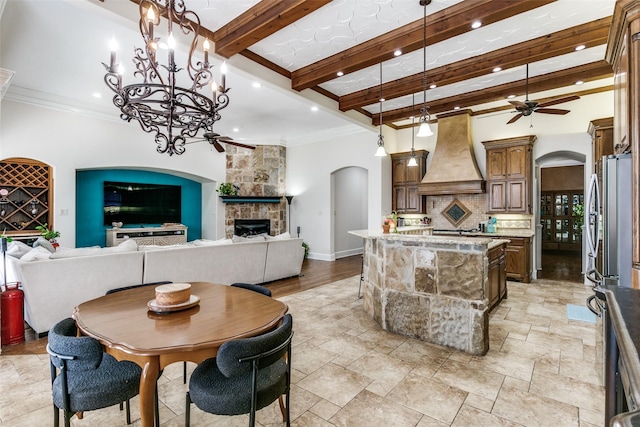 dining room featuring ceiling fan with notable chandelier, ornamental molding, beamed ceiling, and a fireplace