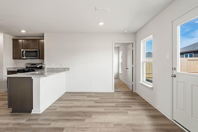 kitchen featuring kitchen peninsula, dark brown cabinetry, light wood-type flooring, light stone countertops, and stainless steel appliances