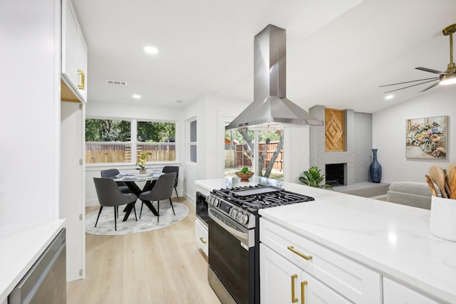 kitchen with white cabinets, gas stove, a brick fireplace, island range hood, and ceiling fan