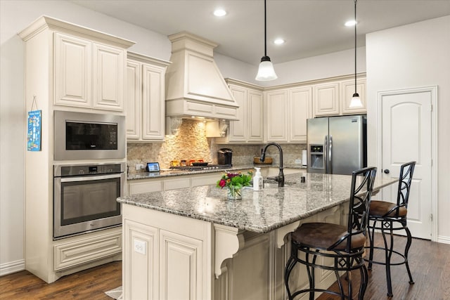 kitchen with stainless steel appliances, decorative backsplash, premium range hood, hanging light fixtures, and a kitchen island with sink