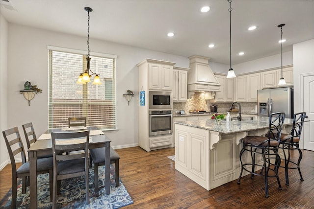 kitchen with decorative light fixtures, custom exhaust hood, a kitchen island with sink, light stone countertops, and stainless steel appliances