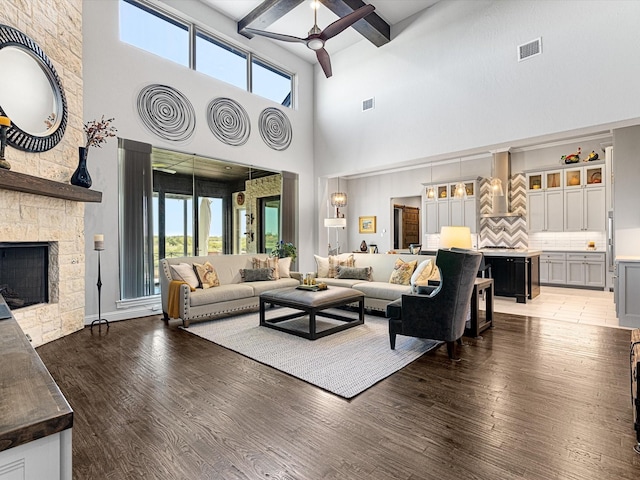 living room with ceiling fan, a towering ceiling, beamed ceiling, a stone fireplace, and dark hardwood / wood-style flooring