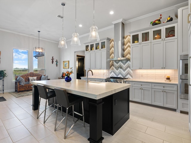 kitchen featuring tasteful backsplash, a kitchen island with sink, pendant lighting, wall chimney exhaust hood, and sink