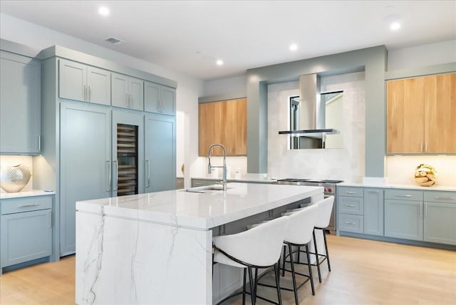 kitchen featuring a kitchen bar, a kitchen island with sink, light wood-type flooring, wall chimney exhaust hood, and sink