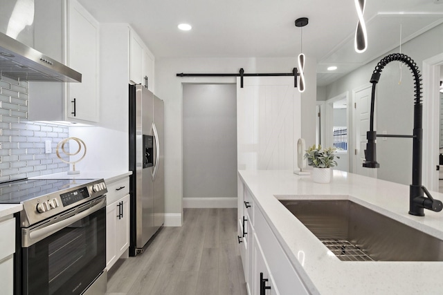 kitchen featuring a barn door, pendant lighting, white cabinetry, stainless steel appliances, and wall chimney exhaust hood