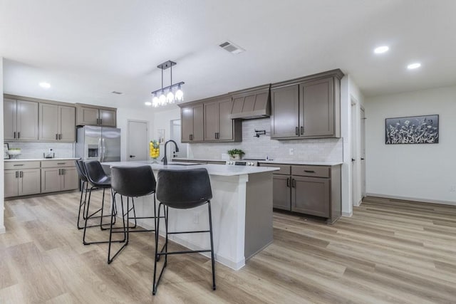 kitchen with wall chimney range hood, pendant lighting, stainless steel refrigerator with ice dispenser, sink, and a kitchen island with sink